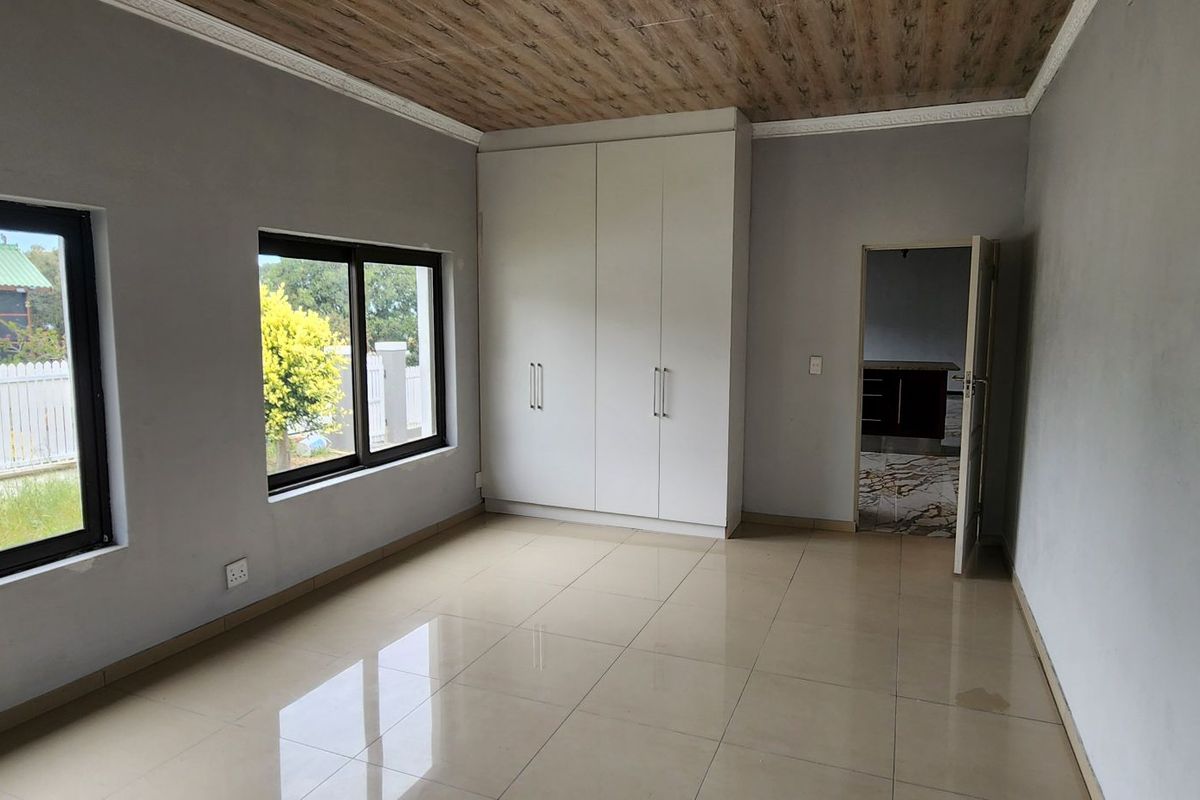 A bright, empty bedroom featuring tiled flooring, built-in cupboards, and modern aluminium window frames with a unique wood-look ceiling finish.