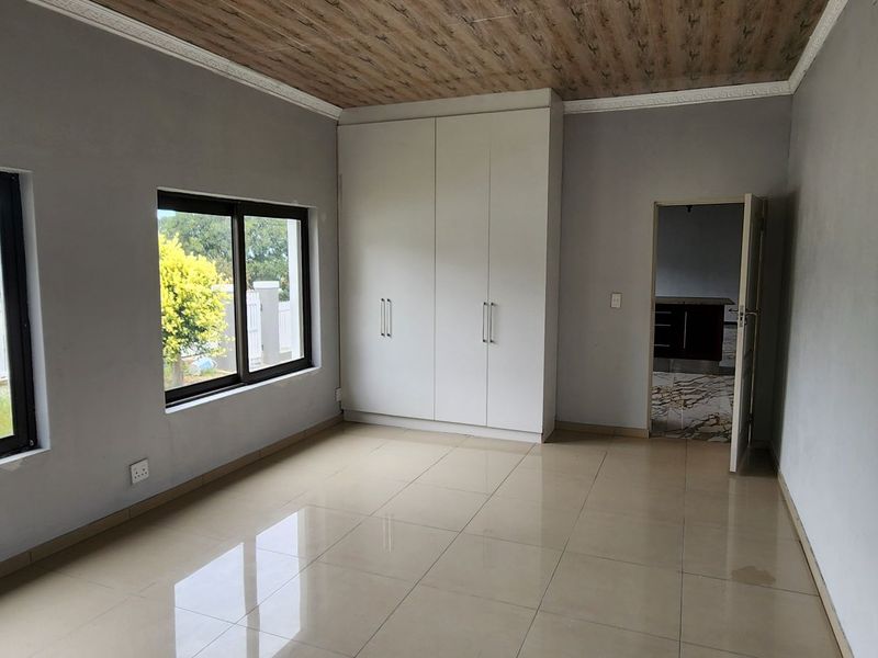 A bright, empty bedroom featuring tiled flooring, built-in cupboards, and modern aluminium window frames with a unique wood-look ceiling finish.