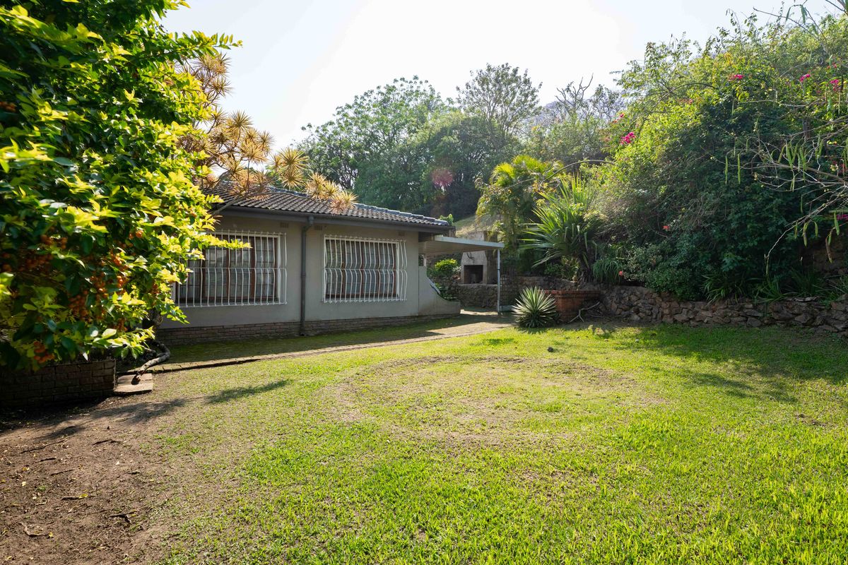 A rear garden view featuring a lawn, paved walkway, and a built-in braai area adjacent to the house with security bars on the windows.