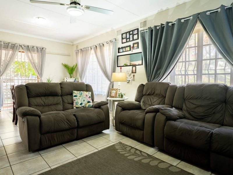 A comfortable living room featuring tiled flooring, a ceiling fan, and large windows with security bars, providing access to the outdoor area.