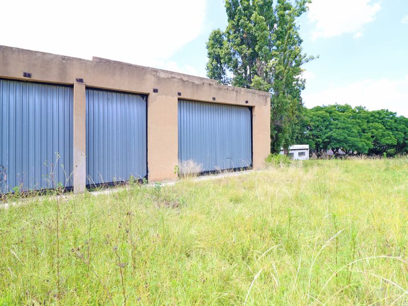 A detached triple garage structure with metal roller doors, situated on a large, overgrown plot of land.