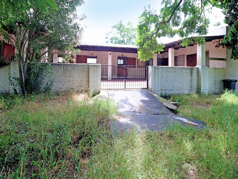 Rear exterior view featuring a covered patio area, concrete boundary wall with metal gate, and an overgrown garden requiring significant landscaping and maintenance.