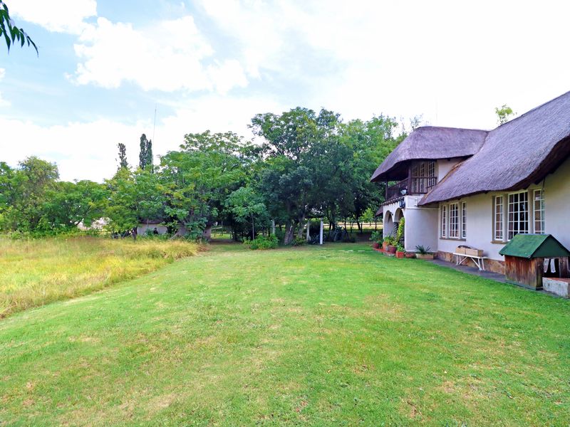 Spacious rear garden featuring a large lawn, mature trees, and a traditional thatched-roof house with a balcony and arched patio area.