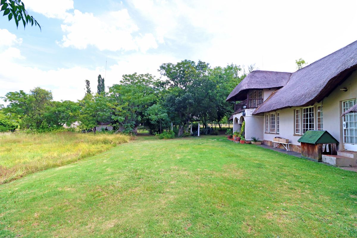 Spacious rear garden featuring a large lawn, mature trees, and a traditional thatched-roof house with a balcony and arched patio area.