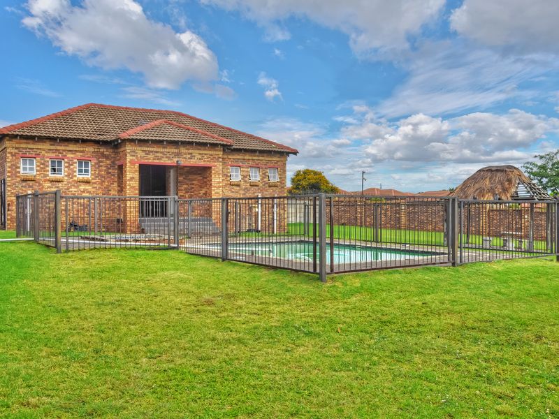 Rear view of the property featuring a brick exterior, a fenced swimming pool, and a thatched lapa set within a spacious garden.