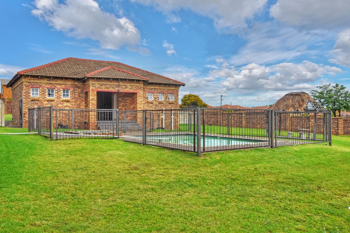 Rear view of the property featuring a brick exterior, a fenced swimming pool, and a thatched lapa set within a spacious garden.
