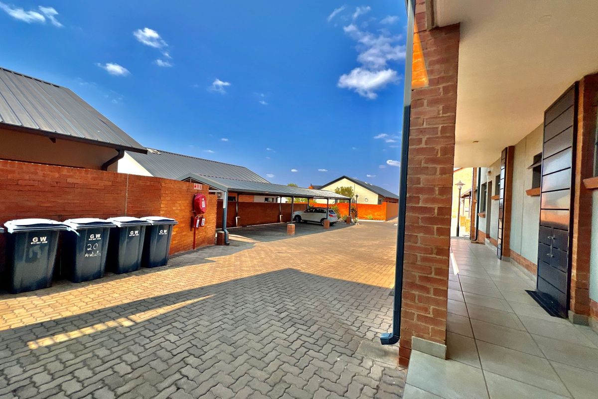 Exterior view of the apartment complex featuring paved driveway, covered parking bays, brick boundary walls, and a tiled walkway.