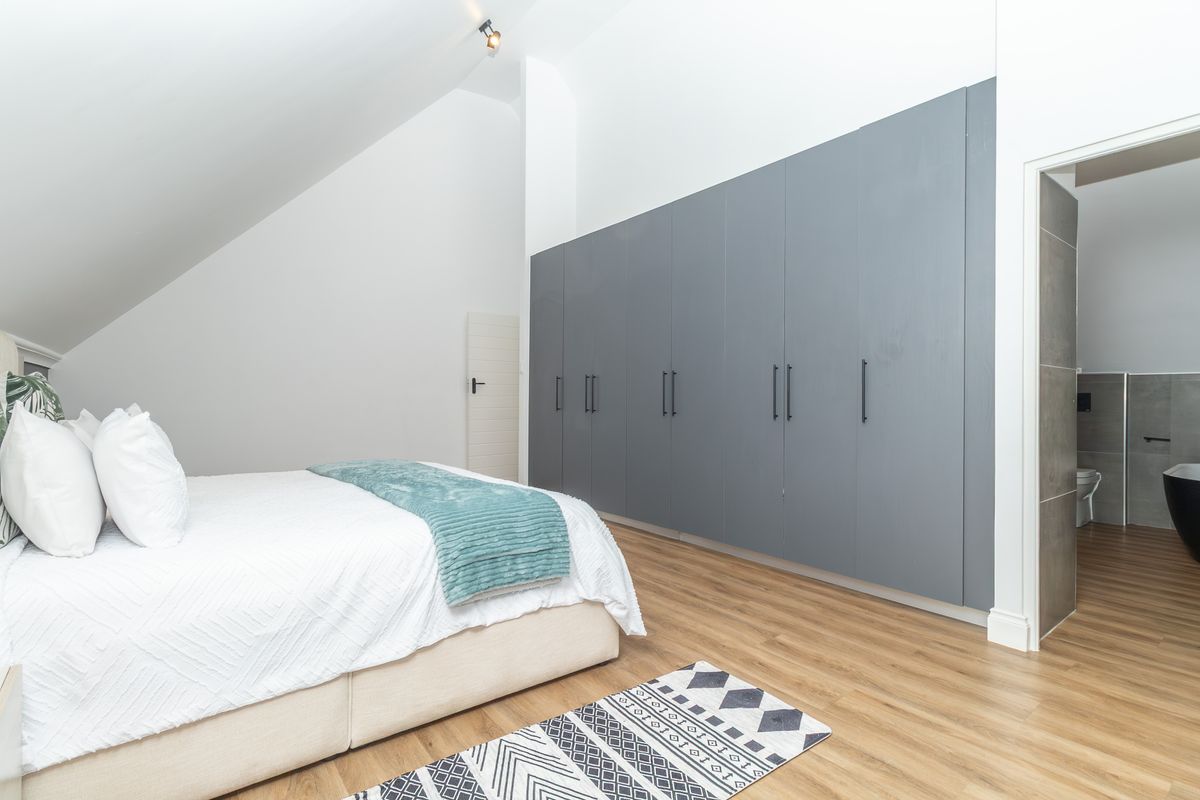 A modern bedroom featuring a large wall-to-wall built-in wardrobe with grey cabinetry, vaulted ceilings, and direct access to an en-suite bathroom.