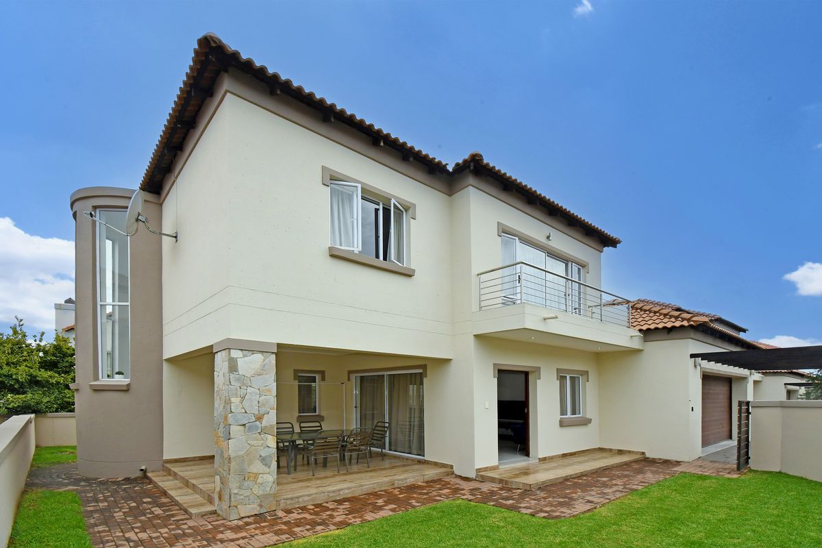 Rear view of the double-story house featuring a covered patio, a balcony with metal railings, stone-clad architectural detailing, and a well-maintained garden.