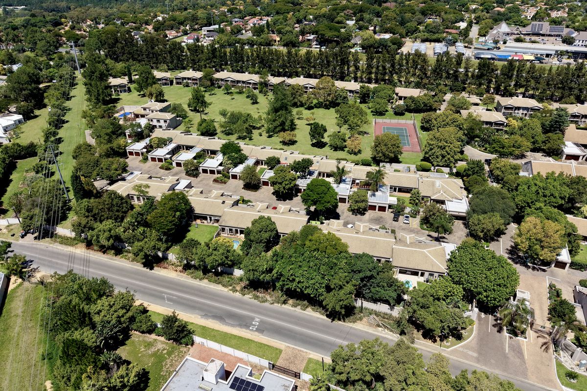 Aerial view of a residential townhouse complex featuring communal gardens, a tennis court, swimming pools, and established trees within a suburban setting.