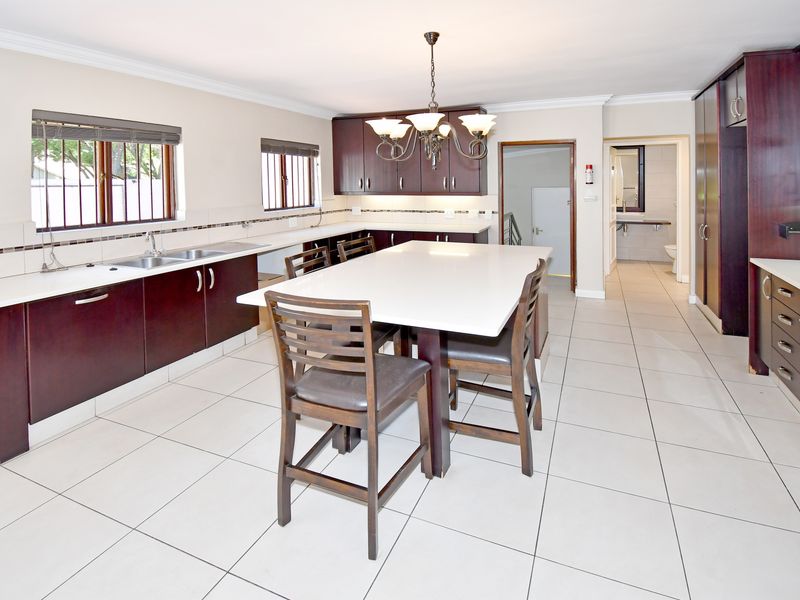 Spacious kitchen featuring dark wood cabinetry, stone countertops, integrated appliances, and tiled flooring, with an adjoining breakfast island and access to a rear hallway.