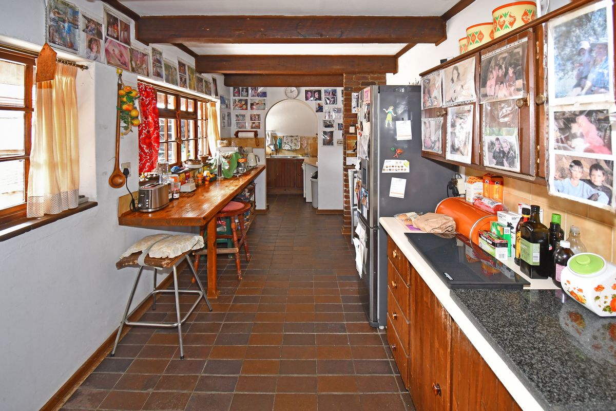 A dated kitchen featuring wooden cabinetry, a breakfast bar, exposed ceiling beams, and terracotta floor tiles, leading through an archway to a scullery area.
