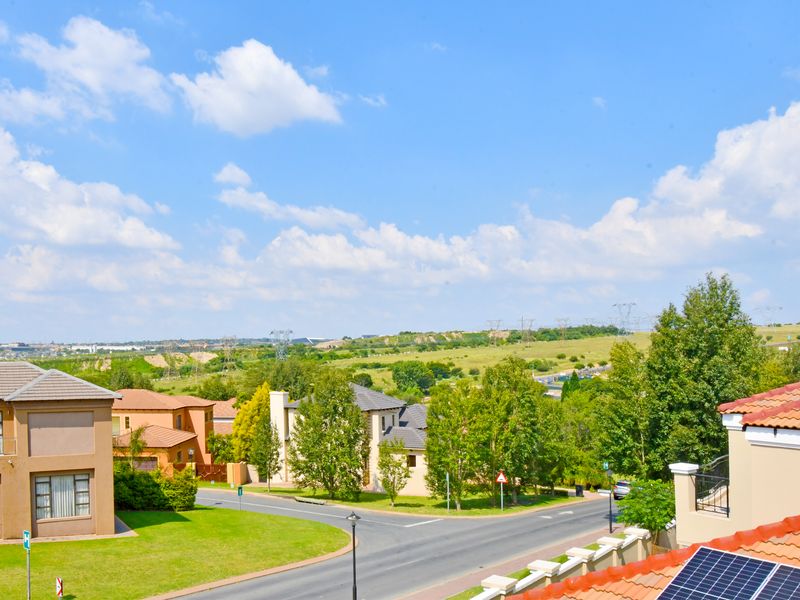 Elevated view overlooking a suburban residential estate with green landscapes, paved roads, and surrounding houses under a clear blue sky.