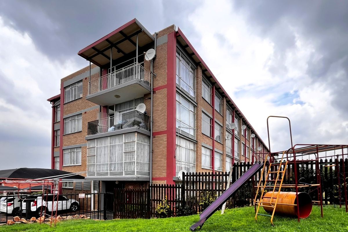 Exterior view of a multi-story brick apartment building featuring balconies, communal playground area, and covered parking facilities.