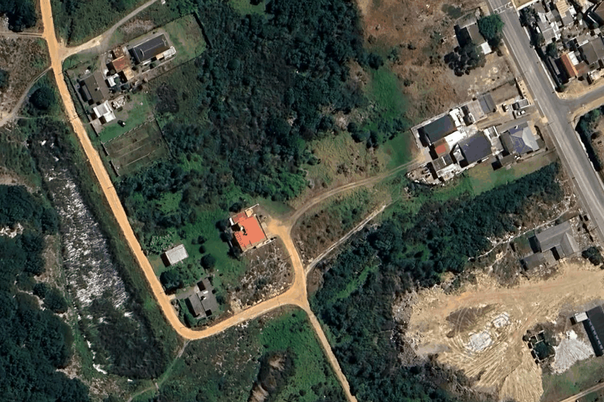 Aerial view of a residential land plot featuring existing structures, surrounding vegetation, and unpaved access roads in a semi-developed area.