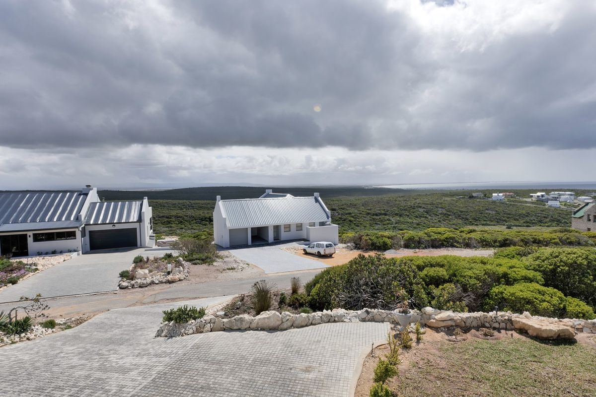 An elevated aerial view of a coastal residential property featuring modern homes with metal roofs, paved driveways, and natural fynbos landscaping.
