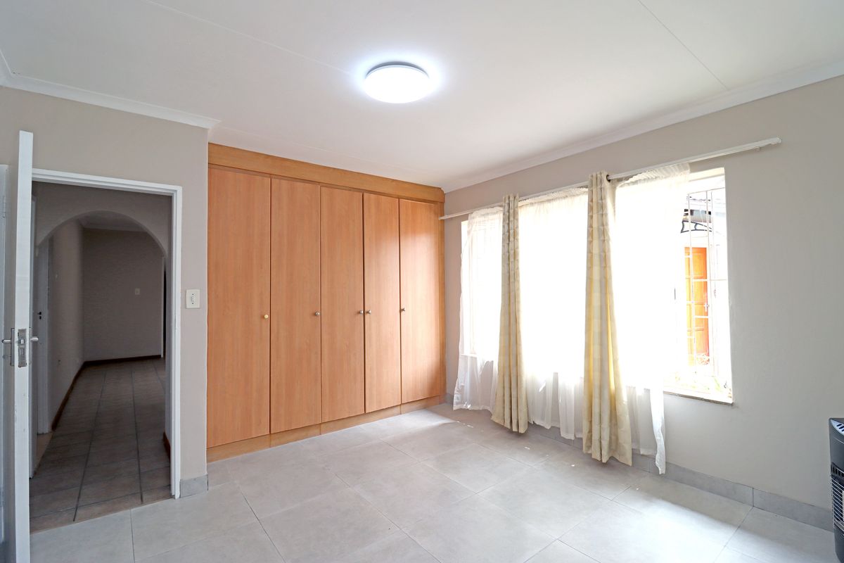 A bright, empty bedroom featuring neutral tiled flooring, built-in wooden cupboards, and large windows with security bars.