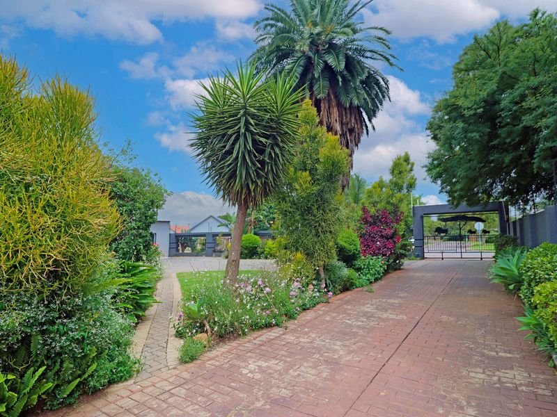 A well-maintained front entrance featuring a paved driveway, established garden landscaping, and a secure metal gate leading to the property.