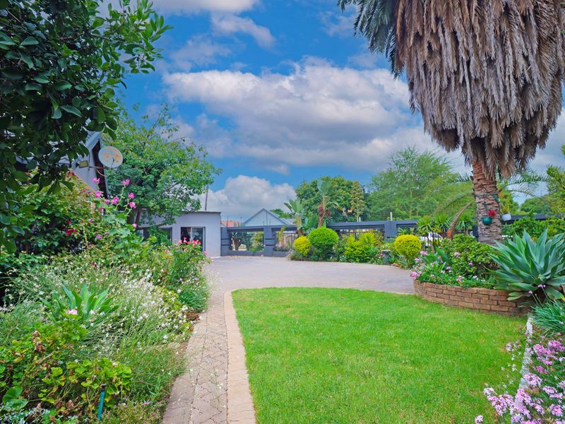 A well-maintained front garden featuring a paved driveway, lush lawn, established trees, and brick-edged garden beds leading to the property entrance.