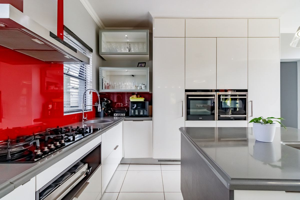 Modern kitchen featuring high-gloss cabinetry, stone countertops, a central island, and integrated appliances with a striking red glass splashback.