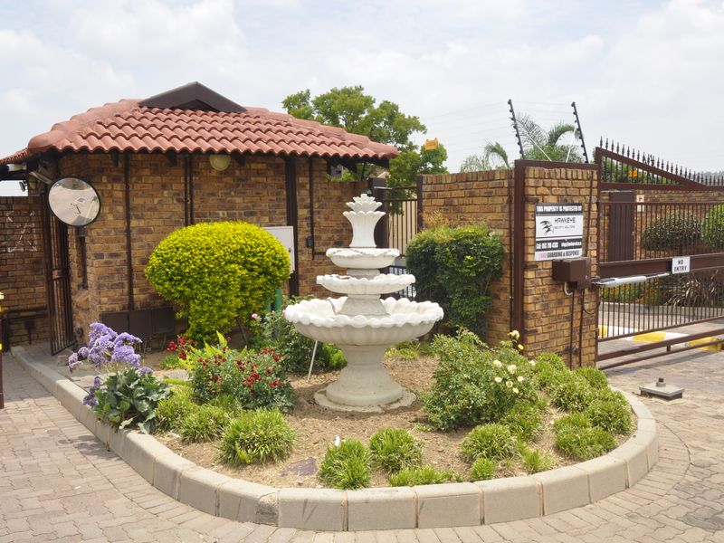 Secure gated entrance to the townhouse complex featuring a brick guard house, automated sliding gate, electric fencing, and a landscaped garden with a central fountain.
