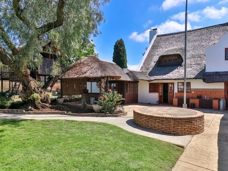 Rear garden area featuring a thatched roof house, paved patio, brick braai, and a mature garden with a treehouse structure.
