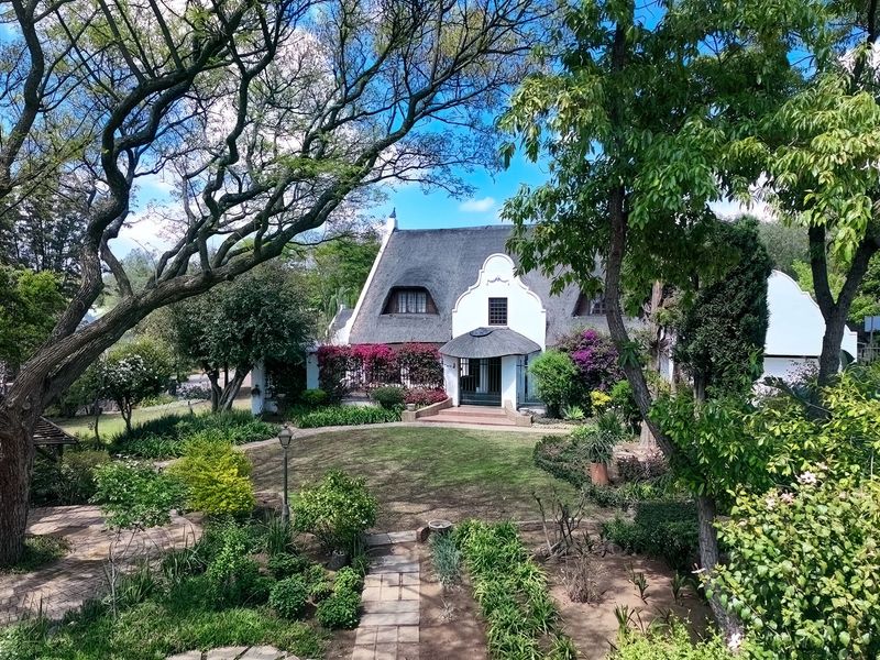 A picturesque view of the rear garden featuring a charming Cape Dutch style home with a traditional thatched roof and established landscaping.
