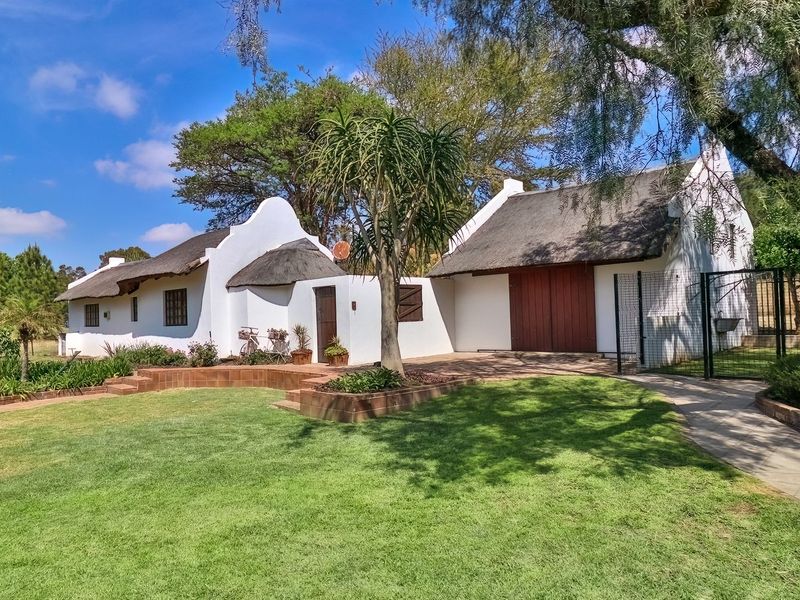 A charming Cape Dutch style cottage featuring a traditional thatched roof, white rendered walls, and a well-maintained garden with brick paved pathways.