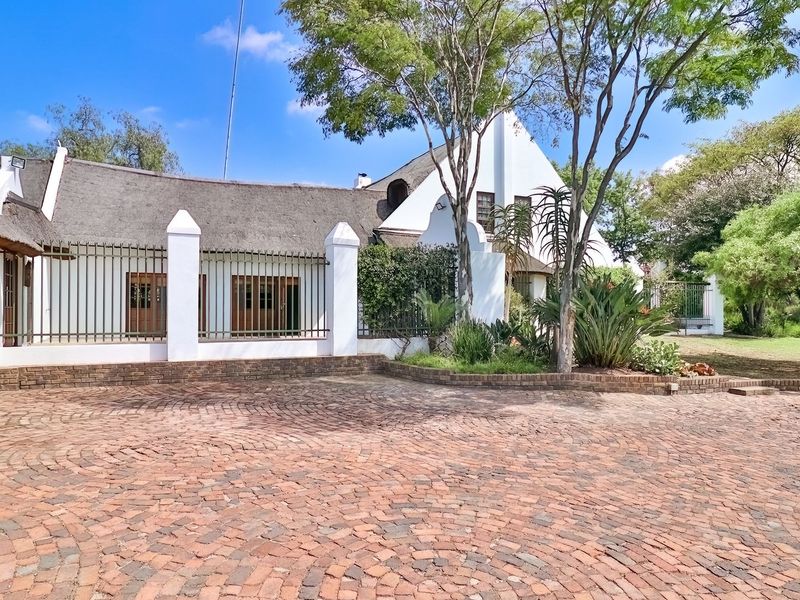 A well-maintained front exterior view featuring a traditional thatched roof, brick paved driveway, and secure metal fencing with masonry pillars.
