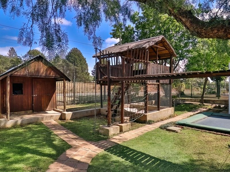 A well-maintained garden featuring a wooden play structure with a thatched roof, a separate wooden shed, and an in-ground trampoline.
