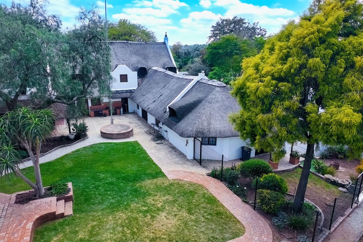 Aerial view of a property featuring a traditional thatched roof house, expansive lawn, brick paved walkways, and a built-in outdoor braai area.