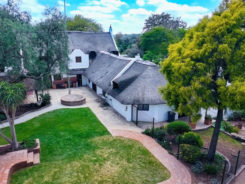 Aerial view of a property featuring a traditional thatched roof house, expansive lawn, brick paved walkways, and a built-in outdoor braai area.