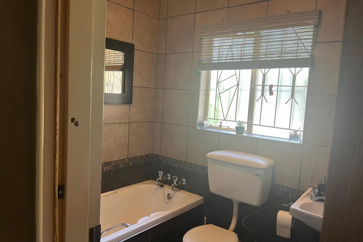 A functional bathroom featuring a built-in bathtub, pedestal washbasin, and toilet, finished with neutral ceramic wall tiling and security bars on the window.