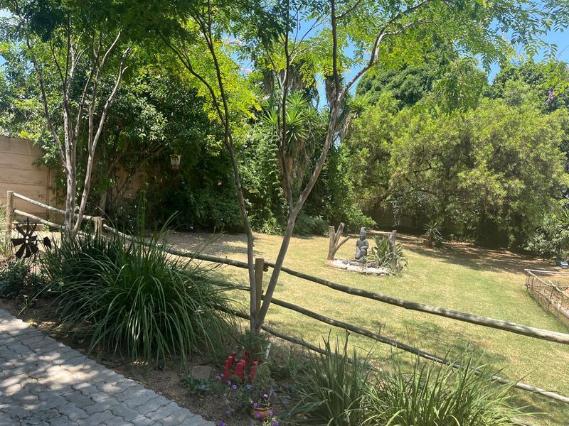 A lush, private garden area featuring a well-maintained lawn, mature trees, and a rustic timber post and rail fence bordering a paved walkway.