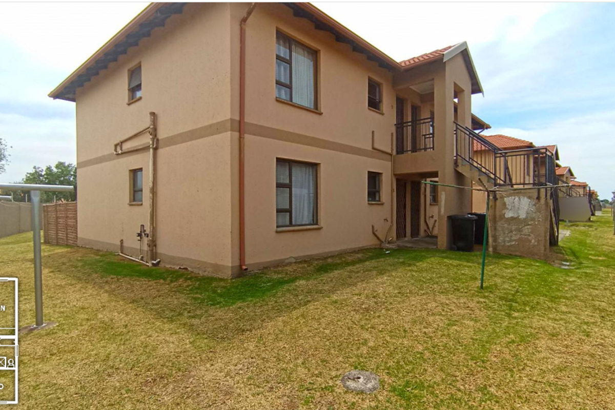 Rear view of a two-story apartment building featuring a balcony, external staircase, and surrounding garden area with perimeter wall and electric fencing.