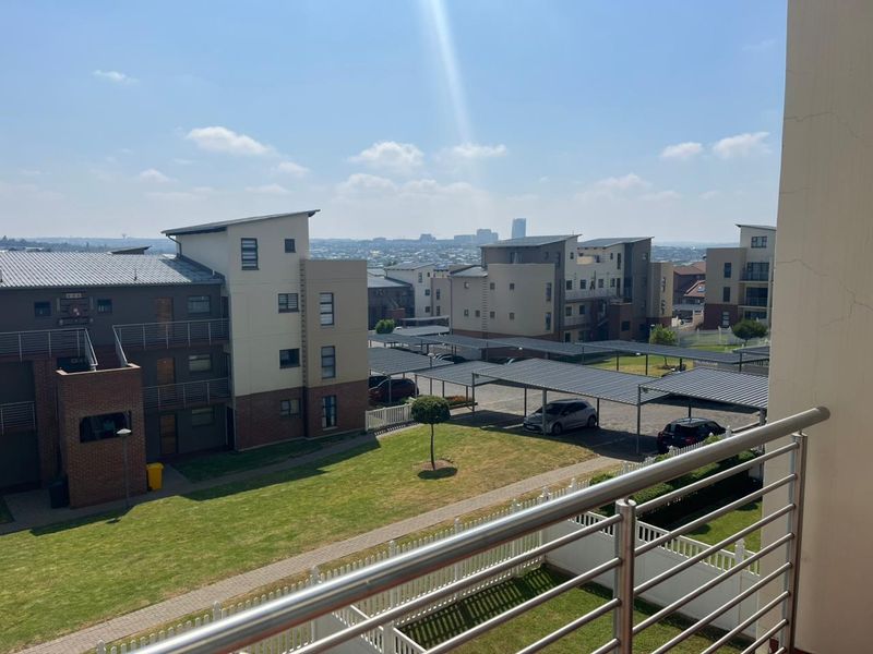 Elevated view from a balcony overlooking the apartment complex grounds, featuring covered parking bays, manicured lawns, and surrounding residential buildings.