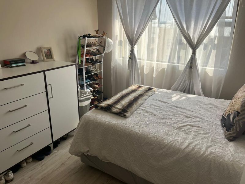 A well-lit bedroom featuring light-toned laminate flooring and a large window with sheer curtains, providing a bright and comfortable living space.