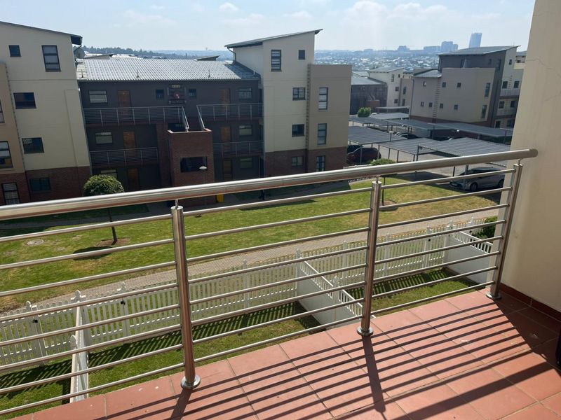 A private balcony featuring a stainless steel balustrade and tiled flooring, overlooking the apartment complex grounds and surrounding neighborhood.