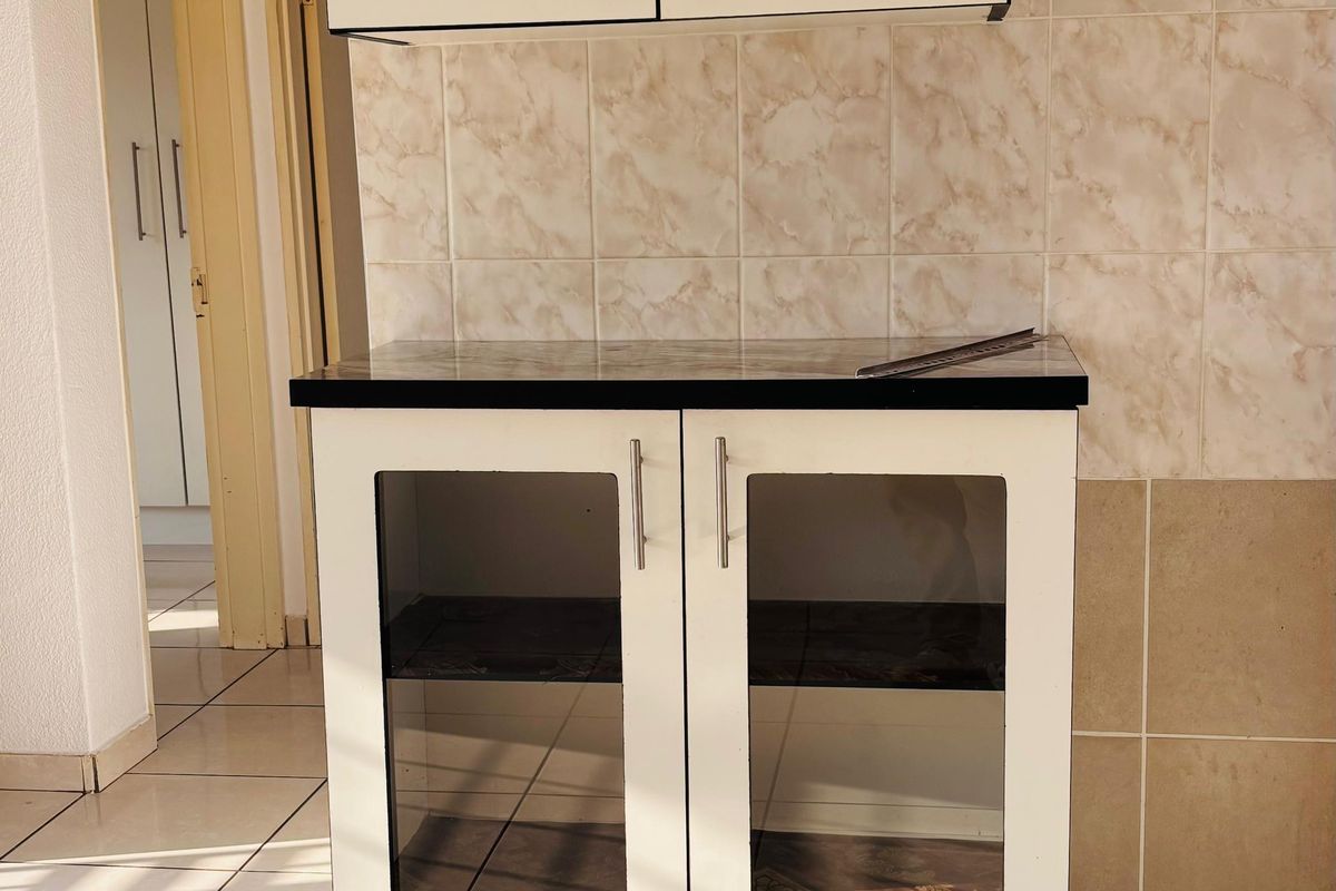A compact kitchen area featuring built-in white cabinetry with glass-fronted lower doors, a dark stone countertop, and neutral tiled splashbacks and flooring.