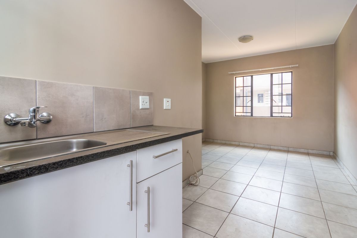 A clean, empty kitchen area featuring tiled flooring, built-in cabinetry with a stainless steel sink, and a window with security bars.