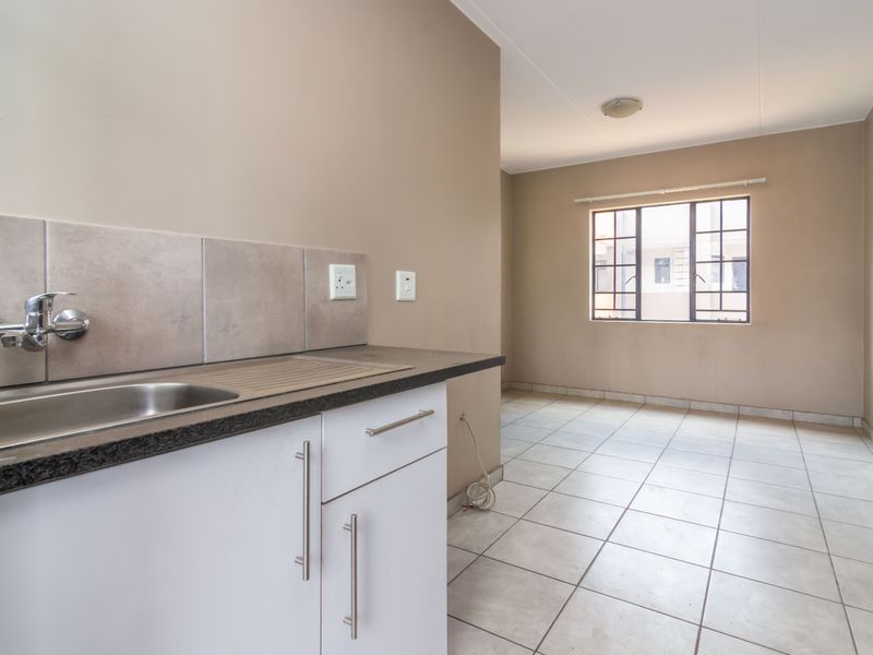 A clean, empty kitchen area featuring tiled flooring, built-in cabinetry with a stainless steel sink, and a window with security bars.