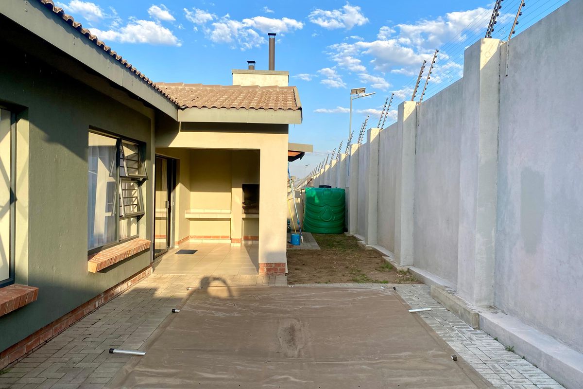 Rear exterior view featuring a covered patio with a built-in braai, paved area, perimeter wall with electric fencing, and a water storage tank.
