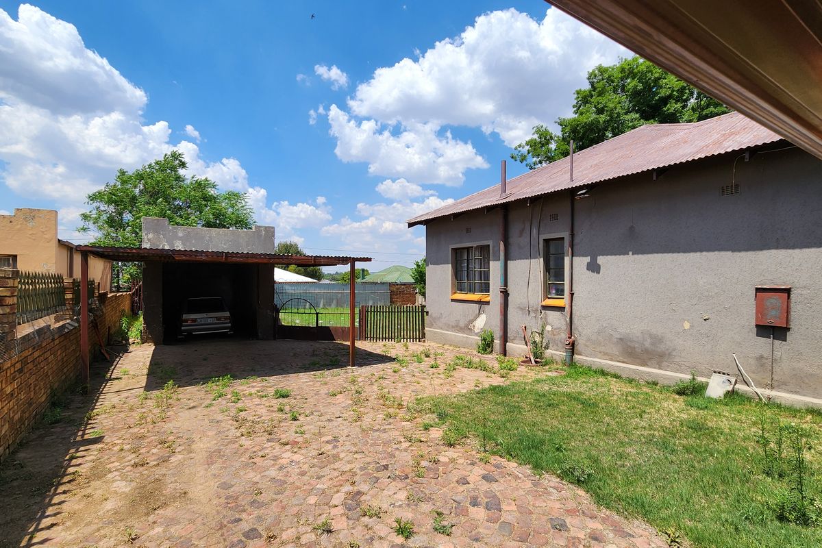 Rear exterior view showing a paved driveway, carport structure, and the back facade of the house with visible wear to the exterior plaster.