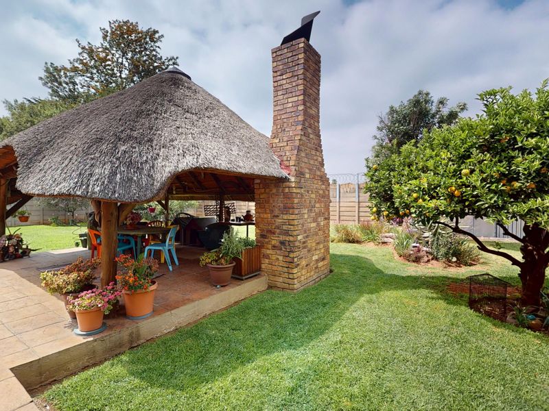 A covered outdoor patio area featuring a traditional thatch roof, brick chimney, and tiled flooring, set within a well-maintained garden.