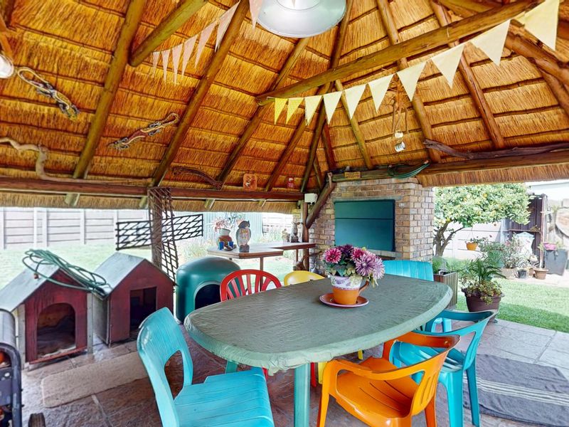 Covered outdoor patio area featuring a traditional thatched roof, exposed timber rafters, and a built-in brick braai, overlooking the rear garden.