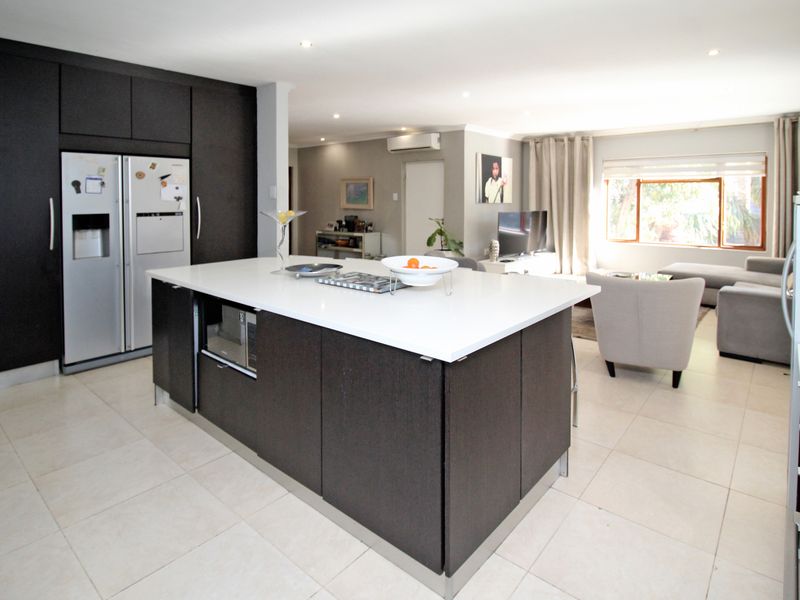 Spacious open-plan kitchen and living area featuring a large central island, modern dark cabinetry, stone countertops, and neutral tiled flooring throughout.
