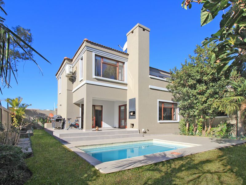 Rear view of a two-story house featuring a tiled patio, built-in braai, and a swimming pool set within a private garden.