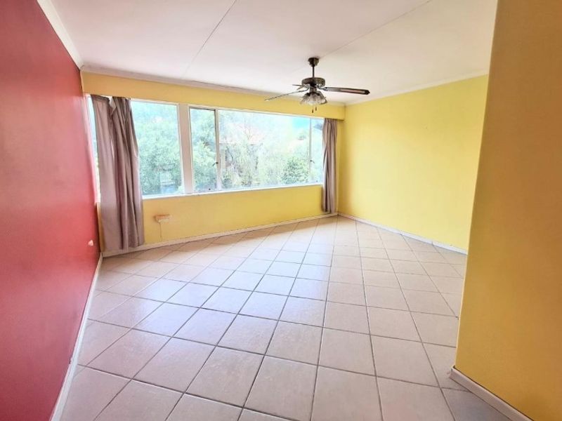 A spacious, empty living room featuring tiled flooring, a ceiling fan, and large windows providing natural light.