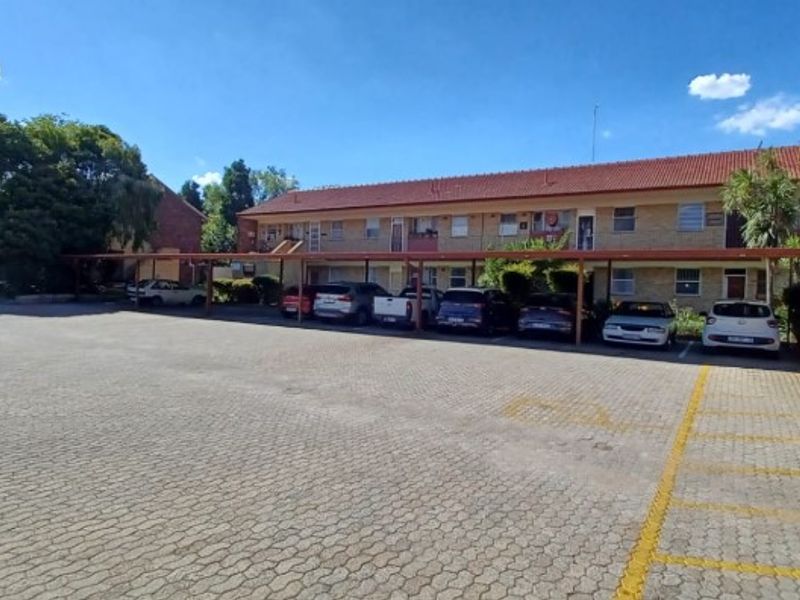 Exterior view of a multi-story flat complex featuring a paved parking area with covered carports and brick building facade.