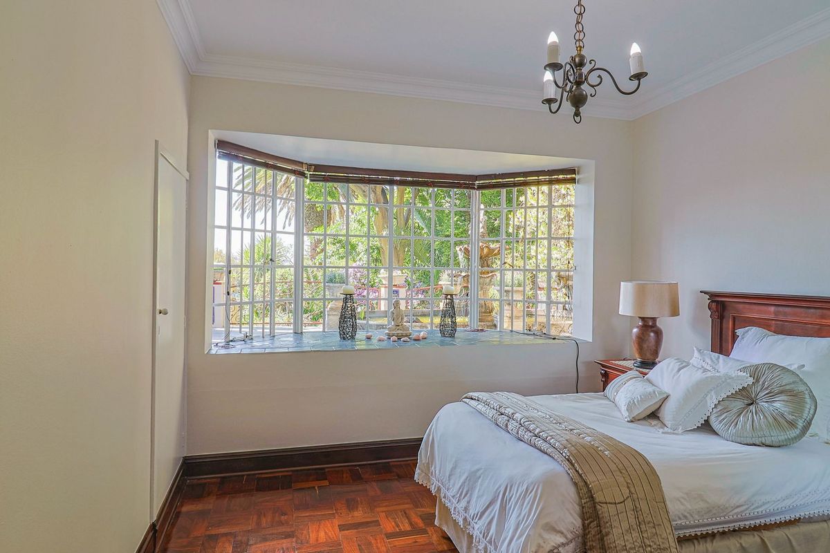 A bright bedroom featuring classic parquet flooring, crown molding, and a large bay window that provides ample natural light and garden views.